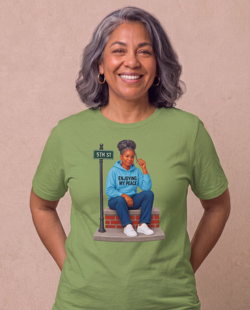 Carmen, a joyful Latina woman in her 50s with wavy dark hair streaked with silver and gold hoops, wearing the Enjoying My Peace Tee in Leaf green with a silver-haired queen relaxing on her front stoop. Hands On 5th St.