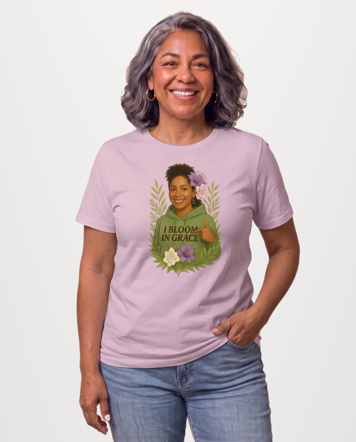 Carmen, a joyful Latina woman in her 50s with wavy dark hair streaked with silver and gold hoops, wearing the I Bloom In Grace Exclusive Tee in Light Violet with a smiling woman in a green hoodie framed by flowers and leaves. Hands On 5th St.