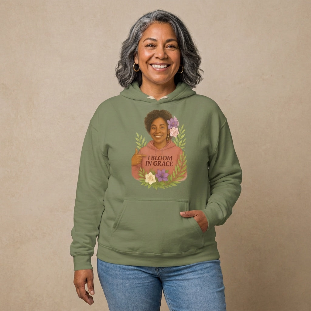 Carmen, a joyful Latina woman in her 50s with wavy dark hair streaked with silver and gold hoops, wearing the I Bloom In Grace Signature Hoodie in Sage with a smiling woman surrounded by flowers and leaves. Hands On 5th St.