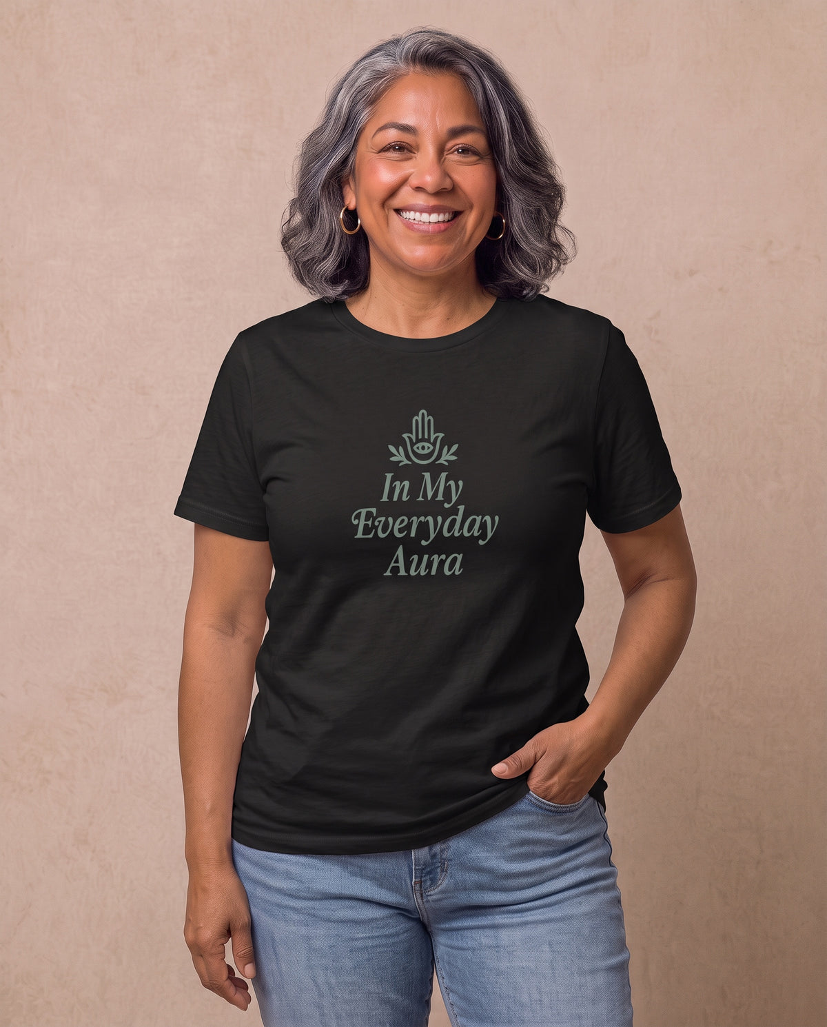 Carmen, a joyful Latina woman in her 50s with wavy dark hair streaked with silver and gold hoops, wearing the In My Everyday Aura Signature Tee in Black with an embellished Hamsa. Hands On 5th St.