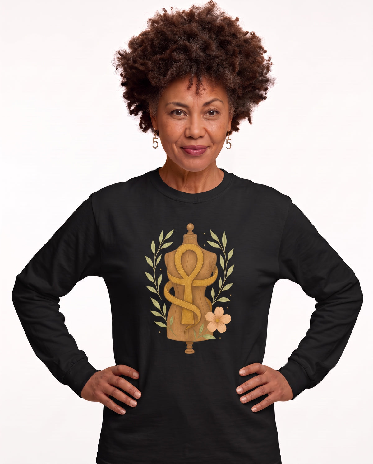 Charmaine, a woman in her 40s with a curly afro and freckles, wearing the Feminine Form of Power Long Sleeve Crewneck Tee in Black with a gold Ankh wrapped around a dress form, laurel leaves, and a peach bloom. Hands On 5th St.