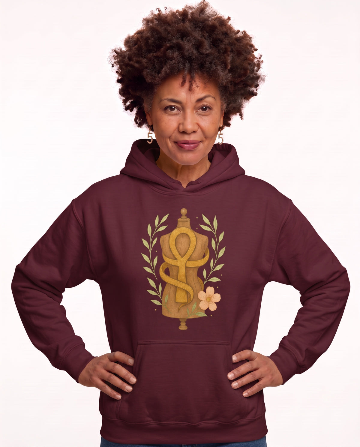 Charmaine, a woman in her 40s with a curly afro and freckles, wearing the Sacred Wine Ankh & Laurel Hoodie in Sacred Wine with a golden Ankh wrapped around a vintage dress form, laurel leaves, and a peach bloom. Hands On 5th St.
