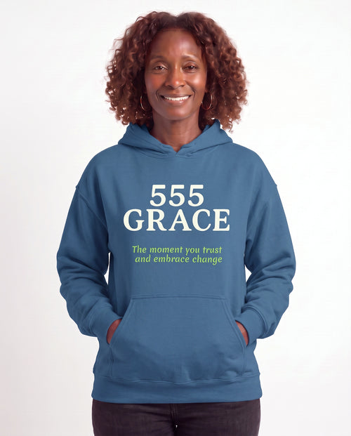 Grace, a Black woman in her 50s with shoulder-length auburn curls and gold hoops, wearing the 555 GRACE Hoodie in Indigo Blue with the words The moment you trust and embrace change. Hands On 5th St.