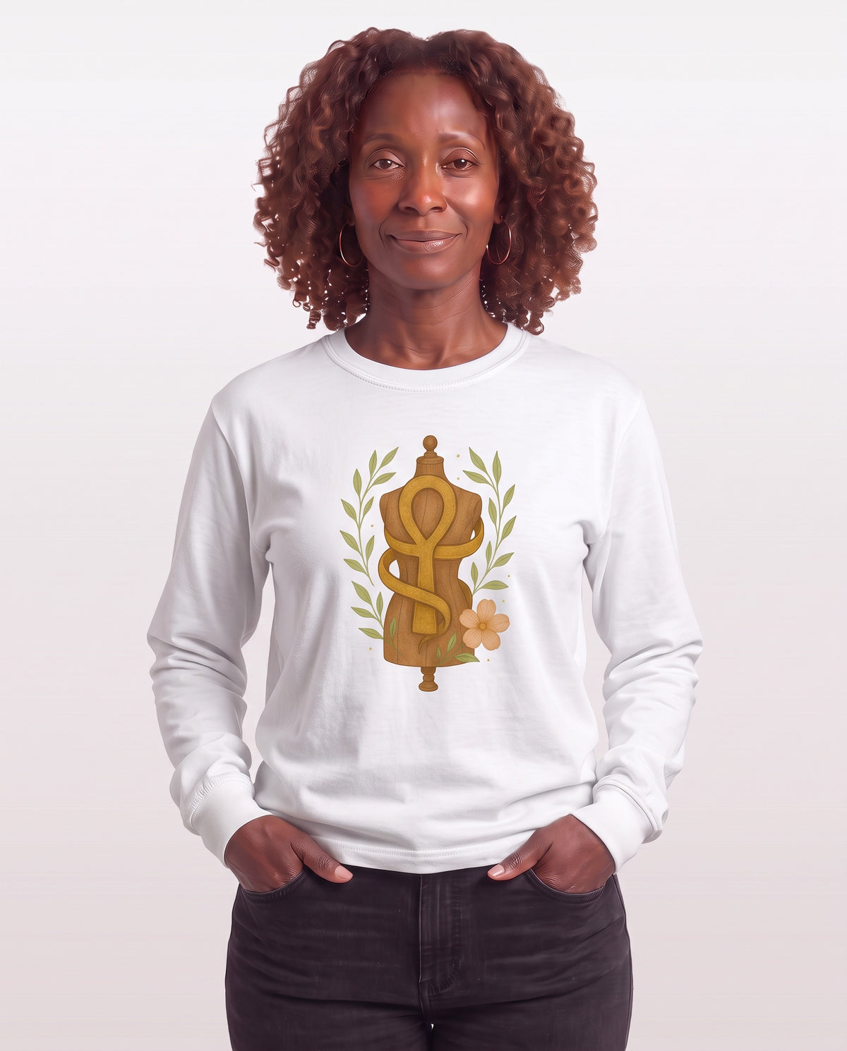 Grace, a Black woman in her 50s with shoulder-length auburn curls and gold hoops, wearing the Feminine Form of Power Long Sleeve Crewneck Tee in White with a gold Ankh wrapped around a dress form, laurel leaves, and a peach bloom. Hands On 5th St.