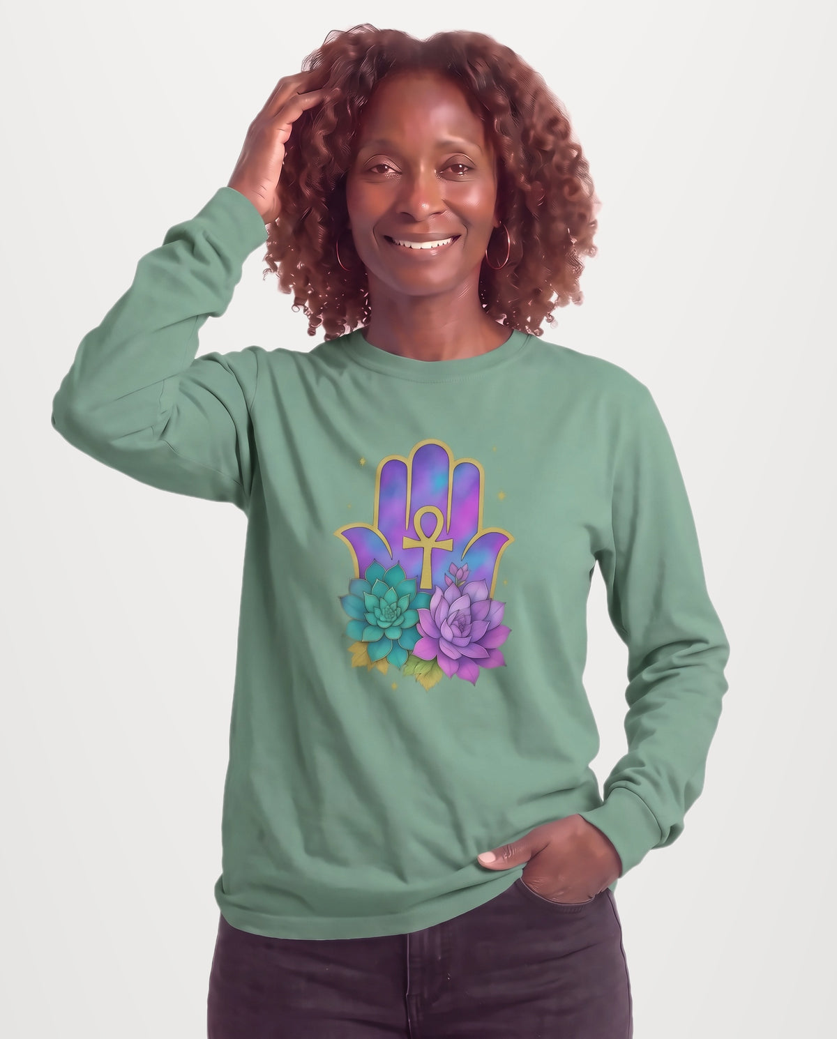 Grace, a Black woman in her 50s with shoulder-length auburn curls and gold hoops, wearing the Hamsa Ankh Bloom Long Sleeve Crewneck Tee in Willow sage green with a gold Ankh, purple Hamsa, and blooming succulents. Hands On 5th St.