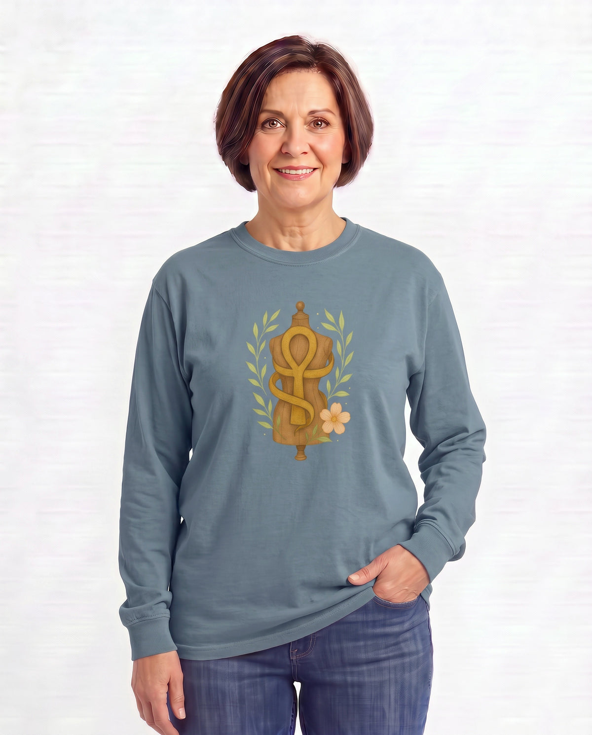 Jillian, a woman in her 50s with a chin-length brown bob, wearing the Feminine Form of Power Long Sleeve Crewneck Tee in Blue Jean with a gold Ankh wrapped around a dress form, laurel leaves, and a peach bloom. Hands On 5th St.