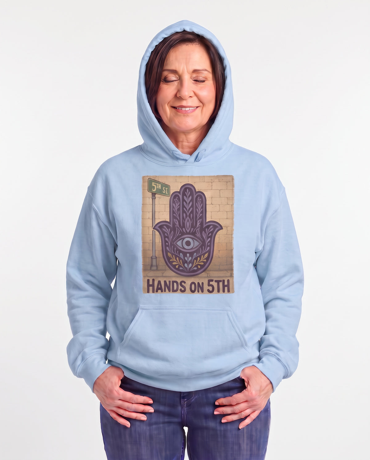 Jillian, a woman in her 50s with a chin-length brown bob, wearing the Hands On 5th St Hoodie in Light Blue with a purple Hamsa beside a 5th St sign. Hands On 5th St.