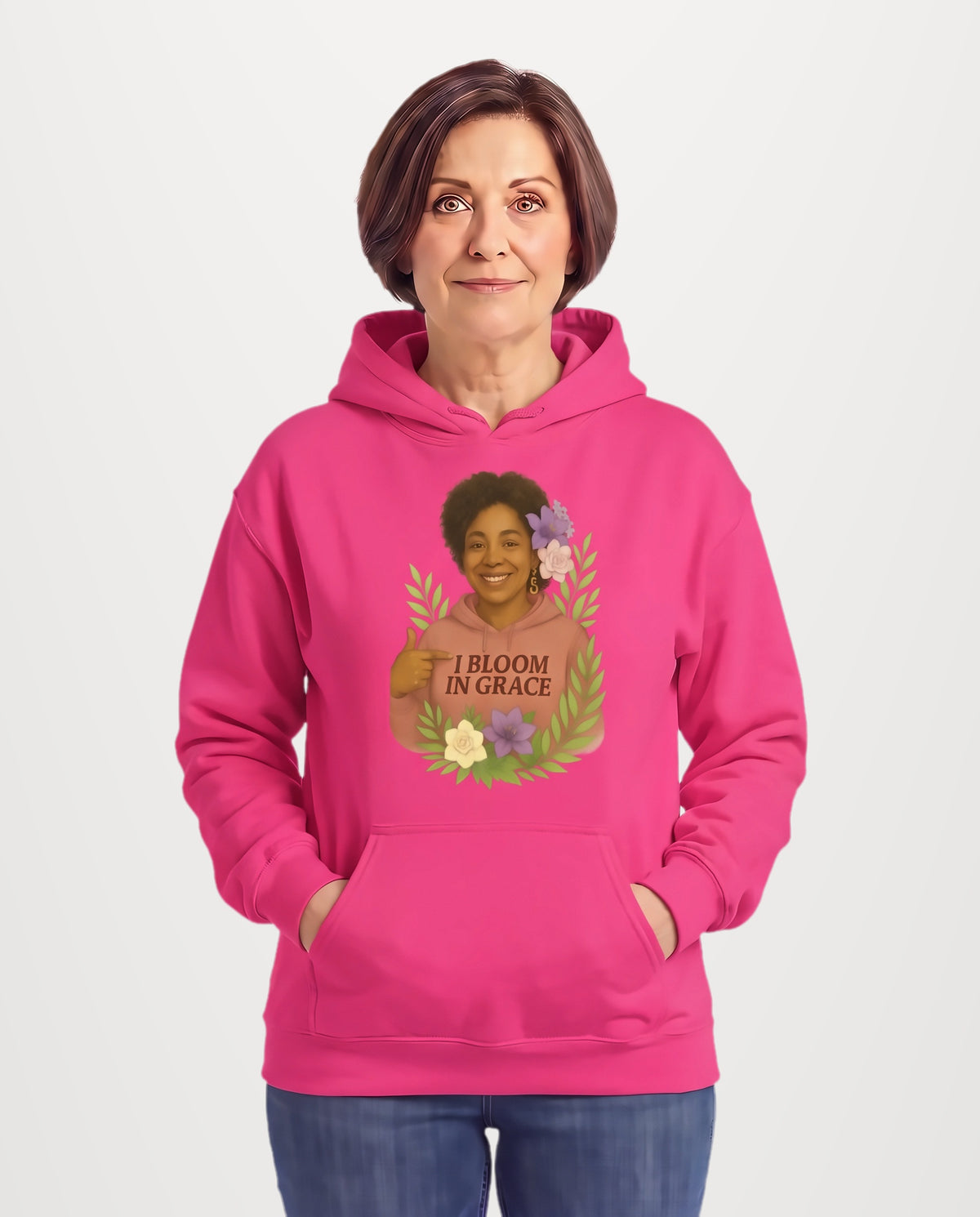 Jillian, a woman in her 50s with a chin-length brown bob, wearing the I Bloom In Grace Signature Hoodie in Sacred Blossom pink, featuring a smiling woman surrounded by flowers and leaves. Hands On 5th St.